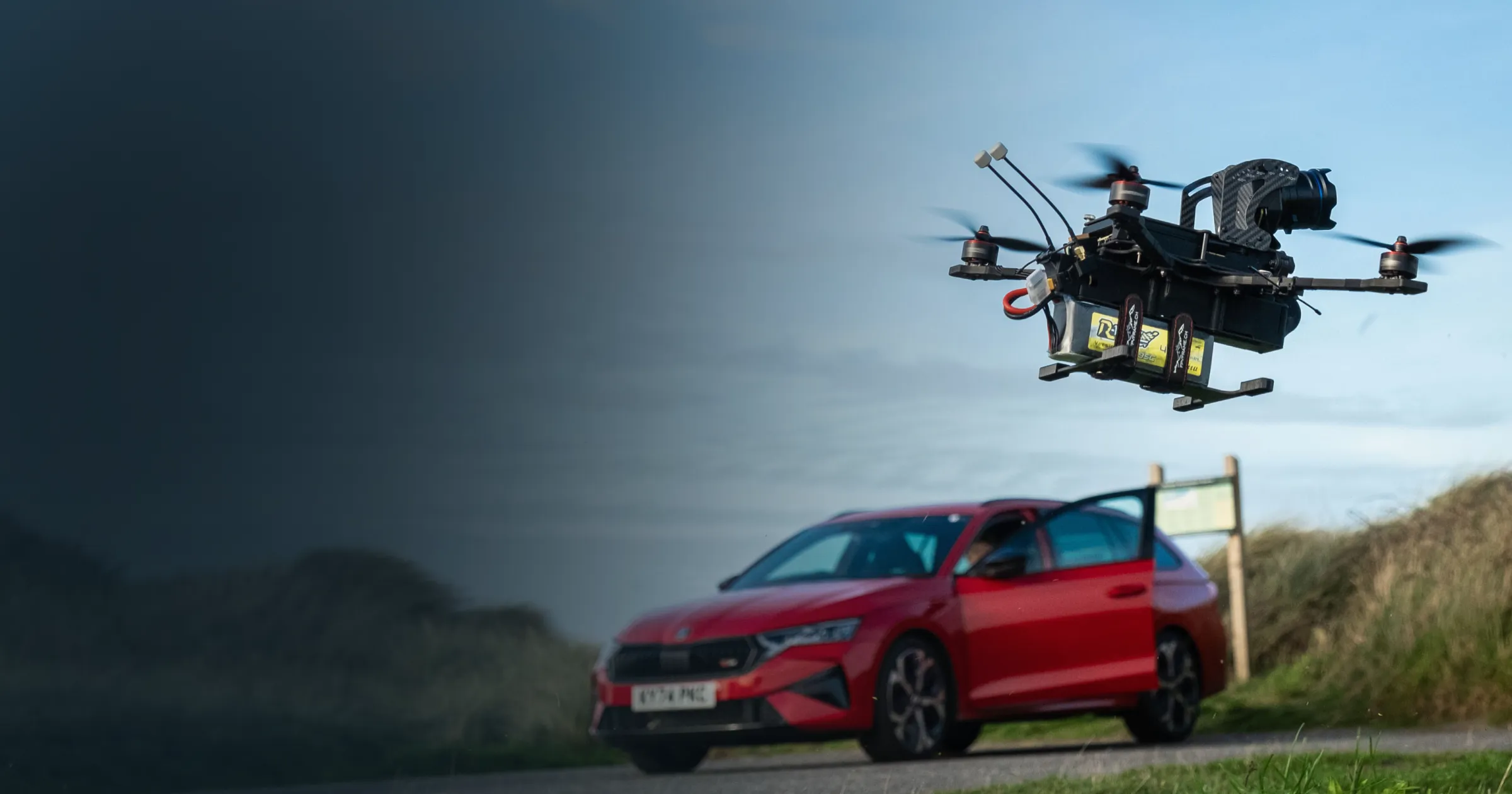 A drone hovers mid-air against a blue sky, with a red Skoda car parked on a rural road in the background. The scene conveys a tech-savvy and adventurous tone.